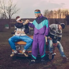 Trzech mężczyzn w strojach w stylu lat 90. siedzi na masce starego samochodu.