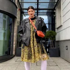 Kobieta w skórzanej ramonesce i kwiecistej sukience, z torebką na ramieniu. Stylizacja nawiązuje do mody z lat 80., przypominając styl prl.