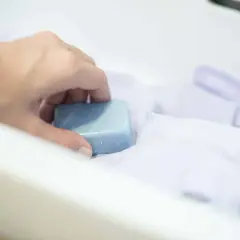 Ręka trzyma kostkę mydła nad białym materiałem. Mydło to skuteczny sposób, czym usunąć klej z tkaniny.