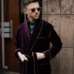 Elegancki mężczyzna w bordowej bonżurce i okularach przeciwsłonecznych, oparty o kamienną ścianę.