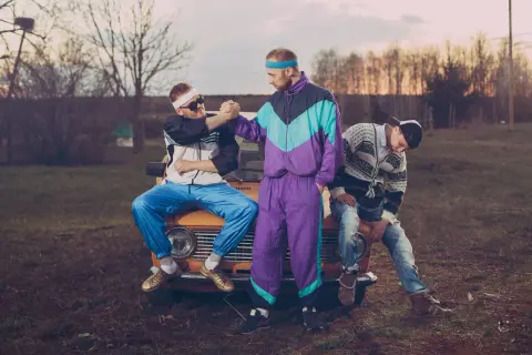 Trzech mężczyzn w strojach w stylu lat 90. siedzi na masce starego samochodu.