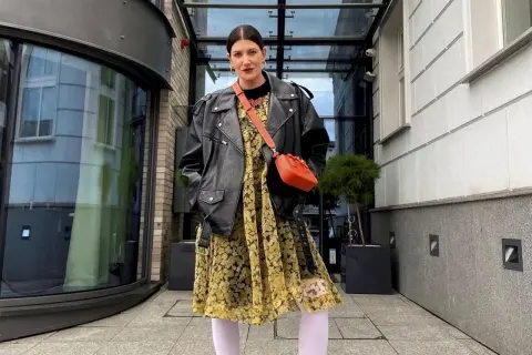 Kobieta w skórzanej ramonesce i kwiecistej sukience, z torebką na ramieniu. Stylizacja nawiązuje do mody z lat 80., przypominając styl prl.