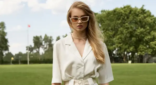 Młoda kobieta w okularach i eleganckim, białym stroju stoi na tle zielonego parku.