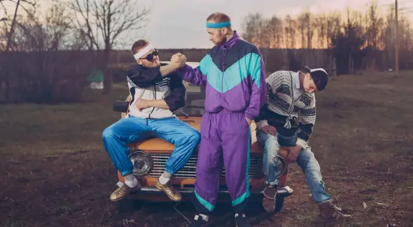Trzech mężczyzn w strojach w stylu lat 90. siedzi na masce starego samochodu.