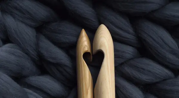 Dwa drewniane szydełka tworzą serce na tle grubiutkiej, ciemnoszarej wełny merino. Co to jest wełna merino? To miękkie włókno idealne do dziergania.