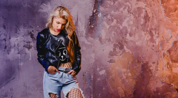 Blondynka w skórzanej kurtce i podartych dżinsach, z siatką na nogach, emanuje **grunge**'owym klimatem przy chropowatej ścianie.