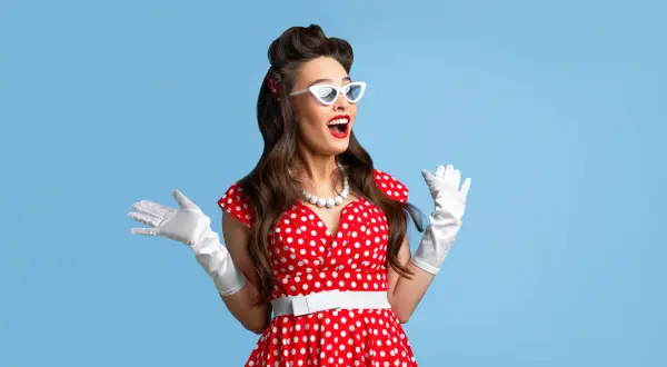 Kobieta w stylu pin-up w czerwonej sukience w grochy, białych rękawiczkach i okularach, z szeroko otwartymi ustami i rękami uniesionymi w geście zaskoczenia.