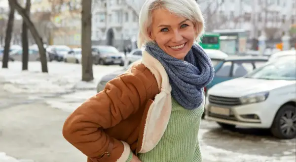 Uśmiechnięta kobieta w brązowym kożuchu i niebieskim szaliku stoi na tle zimowego miasta.