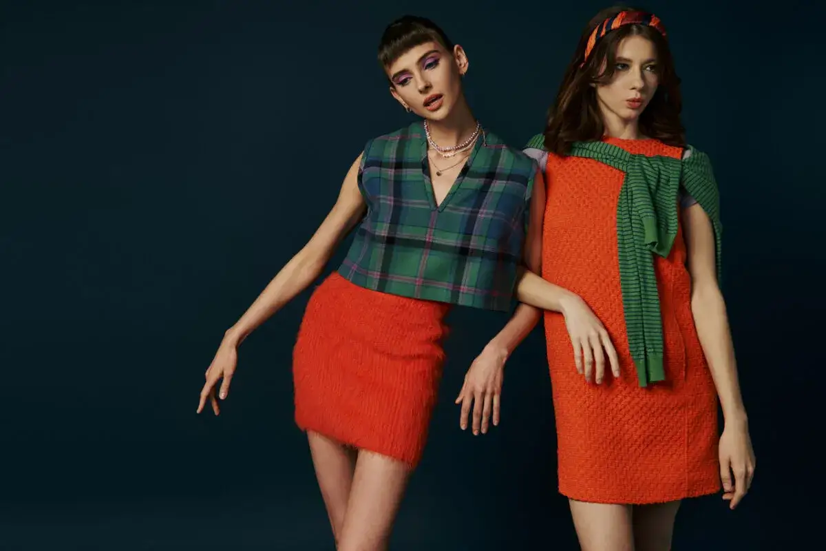 Dwie modelki prezentują stylizacje inspirowane modą lat 60.: krótka, pomarańczowa spódniczka i kraciasty top zestawione z pomarańczową sukienką i zielonym swetrem.