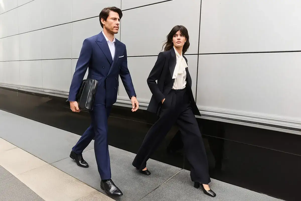 Elegancka para w strojach business formal, mężczyzna w granatowym garniturze, kobieta w czarnym, idą obok siebie.