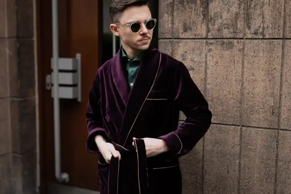 Elegancki mężczyzna w bordowej bonżurce i okularach przeciwsłonecznych, oparty o kamienną ścianę.